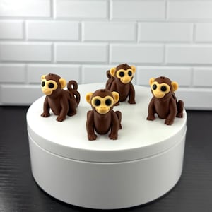 May include: Four brown monkey figurines with tan faces and ears are arranged on a white, circular platform. The monkeys are in a sitting position, with their arms and legs extended. The background is a white brick wall.