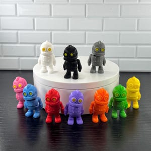 May include: A collection of ten small robot figurines in various solid colors including white, black, gray, pink, blue, red, purple, orange, green, and yellow. Each robot features a rounded head, two yellow circular eyes, and a simple torso design.