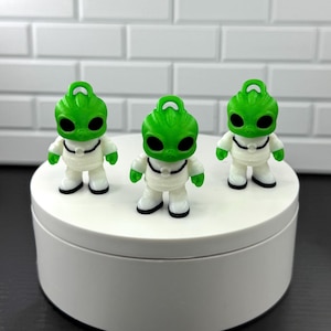 May include: Three small alien figurines. Each figurine has a green head with large black eyes and a small, curved antenna. They wear white suits with black accents and black shoes. The figurines are displayed on a white, circular platform.