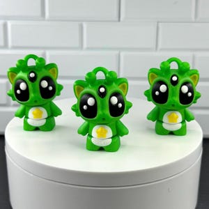 May include: Three small green figurines featuring large black eyes, a third eye on the forehead, and a yellow star emblem on the chest. Each figure has a small loop on top of the head, pointed ears, and a smooth, rounded body design.