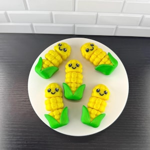 May include: A white plate holds five yellow corn-shaped figurines with smiling faces and green husks. The corn kernels are detailed, and the figurines are arranged in a playful, decorative manner. The background is a white tiled surface.