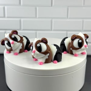 May include: Three small figurines of guinea pigs with brown, white, and black patches. Each figurine features large black eyes, a small pink nose, and pink feet. The guinea pigs are arranged in a group on a smooth, circular white surface.