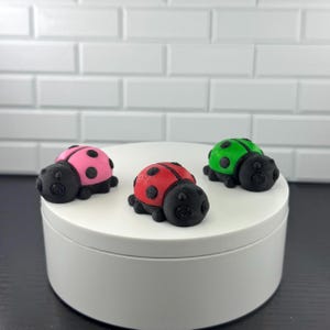 May include: Three colorful ladybug figurines. One is pink with black spots, another is red with black spots, and the third is green with black spots. Each ladybug has a black head and is displayed on a white surface.