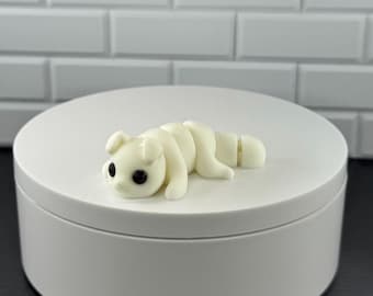 May include: A small, off-white articulated figurine of a creature with rounded ears and two prominent black eyes. The figure has a segmented body design that allows for movement, featuring smooth surfaces and a minimalist, clean aesthetic appearance.