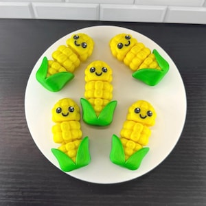 May include: Five small, yellow corn-shaped figurines with green husks at the base. Each corn piece features a smiling face with black eyes and a small mouth. The figurines are arranged in a circular pattern on a white, round surface.