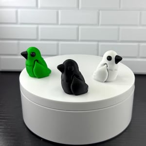 May include: Three small, stylized bird figurines in green, black, and white with black beaks. The birds are displayed on a white, round platform against a white brick background. The figurines appear to be handmade.
