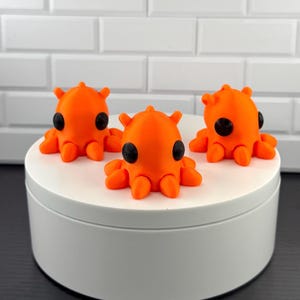 May include: Three bright orange octopus figurines with smooth surfaces and large, round, black eyes. Each figurine features a rounded head with small protrusions and multiple short, curved tentacles extending from the base, arranged in a cluster.