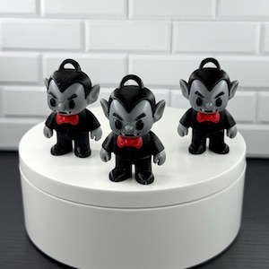 May include: Three small vampire figurines with gray skin, black hair, black suits, and bright red bow ties. Each figure features a small loop at the top of the head, pointed ears, and visible fangs, standing in a group on a circular white platform.