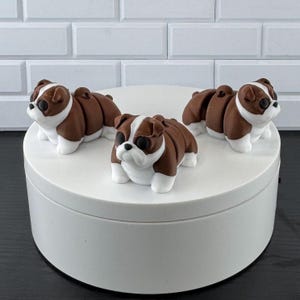 May include: Three small, articulated bulldog figurines featuring a brown and white color scheme. Each dog has a rounded body, dark eyes, and a white muzzle. The figures are positioned on a circular white platform, showcasing their segmented design.