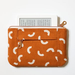 May include: A brown canvas zippered pouch with a white geometric pattern of half circles. The pouch is open and a white e-reader is inside.