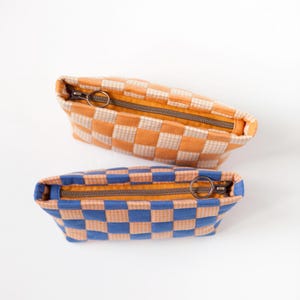 Könnte beinhalten: Zwei kleine, rechteckige Reißverschlusstaschen. Eine ist orange und cremefarben kariert, die andere blau und cremefarben kariert. Beide haben einen braunen Reißverschluss und einen Ringzug. Die Innenseite beider Taschen ist gelb.