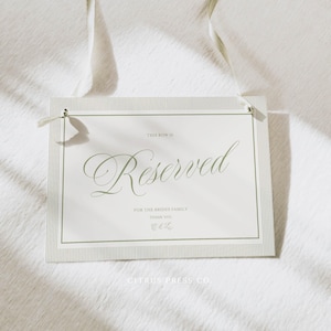 Könnte beinhalten: Rechteckiges Schild mit dem Wort „Reserved“ in eleganter Schrift, grün auf weißem Hintergrund gedruckt. Das Schild enthält Text für die Familie der Braut und ist mit einem weißen Band befestigt. Das Schild ist von Citrus Press Co.