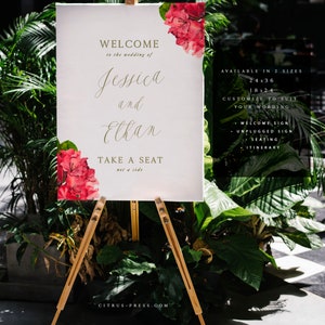 May include: A white welcome sign with gold lettering for a wedding. The sign says "Welcome to the wedding of Jessica and Elkan. Take a seat, not a side." The sign is decorated with red flowers and is on a wooden easel. The sign is available in two sizes: 24x36 inches and 18x24 inches. The sign can be customized to suit your wording. It can be used for a welcome sign, unplugged sign, seating chart, or itinerary.