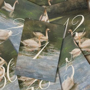 May include: A selection of table number cards with a swan painting. Each card displays a different number in a white script font. The background is a blend of green, blue, and white brushstrokes, creating a water and foliage effect.