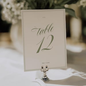 May include: A table number card with the word "Table" in elegant script and the number "12" in a soft green hue. The card is held by a silver metal holder. The text "Cate + Lucas" is below the number.
