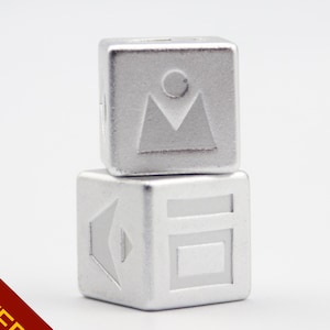 Könnte beinhalten: Zwei gestapelte, silberfarbene Würfel mit geätzten Designs. Der obere Würfel zeigt ein Berg- und Sonnensymbol, während der untere Würfel geometrische Formen zeigt. Vor weißem Hintergrund.