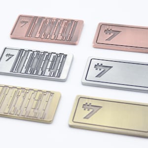 K&ouml;nnte beinhalten: Sechs rechteckige Metall-Namensschilder in verschiedenen Farben: Kupfer, Silber und Gold. Jedes Schild zeigt die Zahl 7 in einer stilisierten Schriftart. Einige Schilder haben ein strukturiertes Design, andere sind glatt.