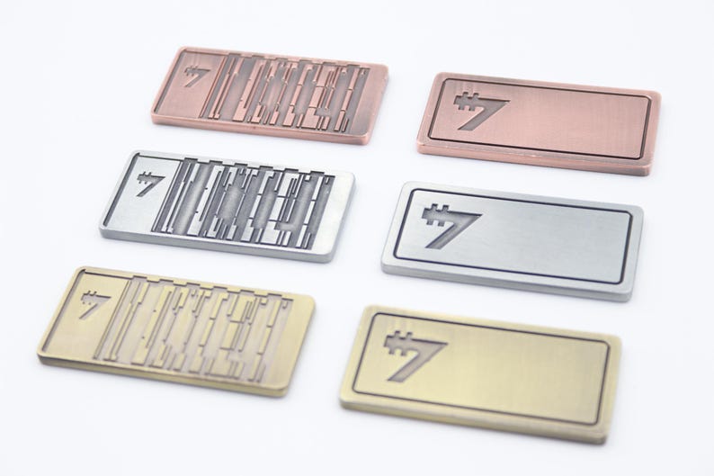 K&ouml;nnte beinhalten: Sechs rechteckige Metall-Namensschilder in verschiedenen Farben: Kupfer, Silber und Gold. Jedes Schild zeigt die Zahl 7 in einer stilisierten Schriftart. Einige Schilder haben ein strukturiertes Design, andere sind glatt.