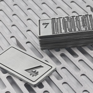 K&ouml;nnte beinhalten: Mehrere rechteckige Metallobjekte mit ge&auml;tzten Designs und der Zahl 7. Ein Objekt liegt separat und zeigt die Zahl 7. Die Objekte sind silberfarben und liegen auf einer perforierten Metalloberfl&auml;che.