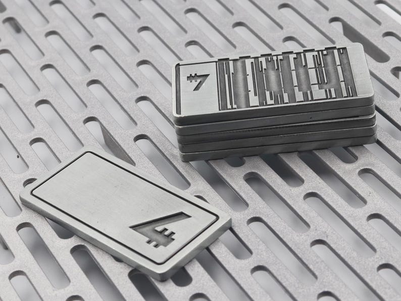 K&ouml;nnte beinhalten: Mehrere rechteckige Metallobjekte mit ge&auml;tzten Designs und der Zahl 7. Ein Objekt liegt separat und zeigt die Zahl 7. Die Objekte sind silberfarben und liegen auf einer perforierten Metalloberfl&auml;che.