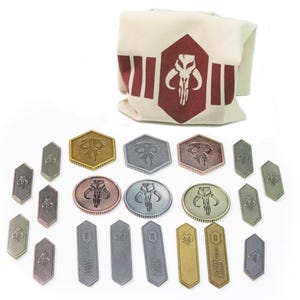 Könnte beinhalten: Eine Sammlung von Metallmünzen und -marken mit einem mandalorianischen Totenkopf-Emblem in verschiedenen Formen und metallischen Ausführungen. Ein Canvas-Beutel mit passendem Emblem ist ebenfalls vorhanden.