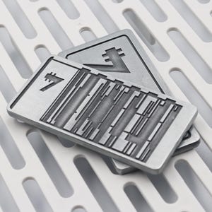 K&ouml;nnte beinhalten: Zwei rechteckige, silberfarbene Metallplaketten mit geometrischen Designs. Eine Plakette liegt auf der anderen, mit einem wei&szlig;en, gemusterten Untergrund. Die Plaketten haben ein strukturiertes Aussehen und weisen abstrakte Symbole auf.