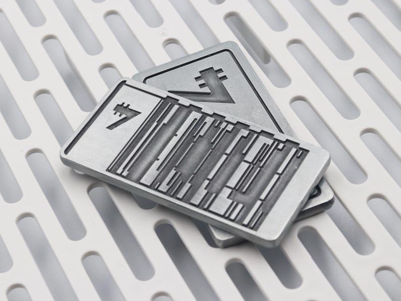 K&ouml;nnte beinhalten: Zwei rechteckige, silberfarbene Metallplaketten mit geometrischen Designs. Eine Plakette liegt auf der anderen, mit einem wei&szlig;en, gemusterten Untergrund. Die Plaketten haben ein strukturiertes Aussehen und weisen abstrakte Symbole auf.