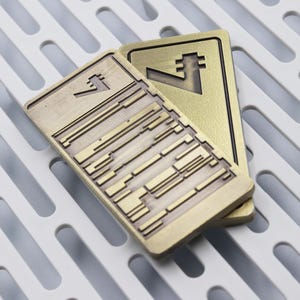 Op de afbeelding: Twee rechthoekige, messingkleurige metalen objecten met geometrische ontwerpen. E&eacute;n heeft een reeks parallelle lijnen en rechthoekige vormen, de andere een gestileerd symbool. De objecten liggen op een wit, sleufvormig oppervlak.