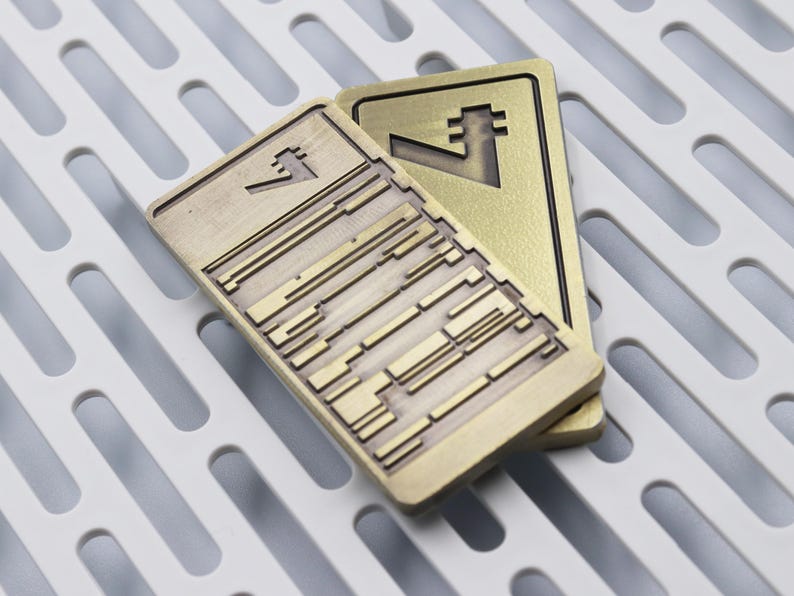 Op de afbeelding: Twee rechthoekige, messingkleurige metalen objecten met geometrische ontwerpen. E&eacute;n heeft een reeks parallelle lijnen en rechthoekige vormen, de andere een gestileerd symbool. De objecten liggen op een wit, sleufvormig oppervlak.