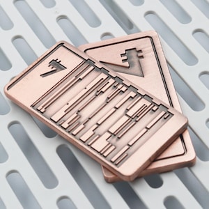 K&ouml;nnte beinhalten: Zwei rechteckige Metallobjekte in Kupferfarbe mit geometrischen Designs. Jedes hat ein einzigartiges Symbol und eine Reihe paralleler Linien und rechteckiger Formen. Die Objekte sind auf einer wei&szlig;en, geschlitzten Oberfl&auml;che gestapelt.