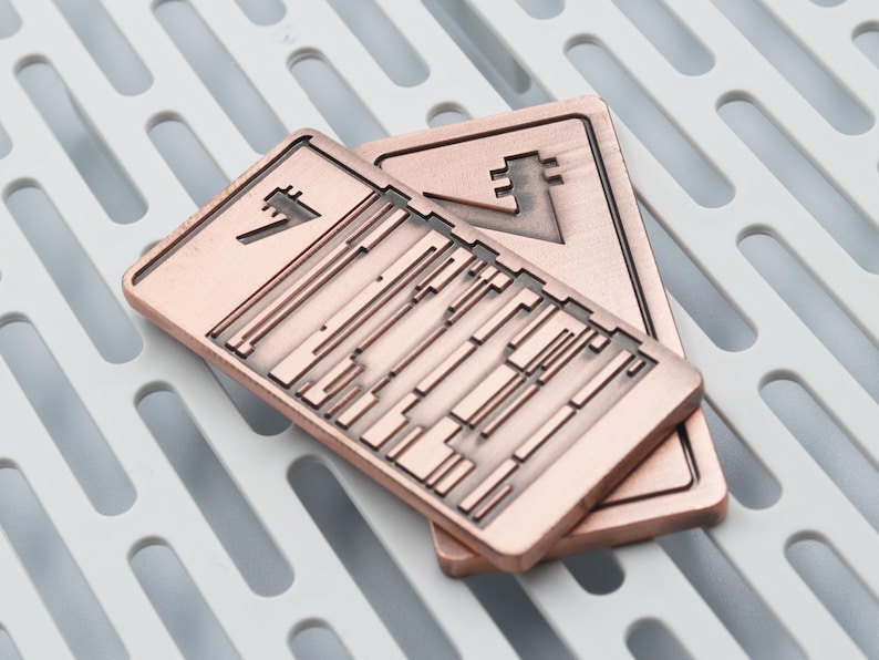 K&ouml;nnte beinhalten: Zwei rechteckige Metallobjekte in Kupferfarbe mit geometrischen Designs. Jedes hat ein einzigartiges Symbol und eine Reihe paralleler Linien und rechteckiger Formen. Die Objekte sind auf einer wei&szlig;en, geschlitzten Oberfl&auml;che gestapelt.