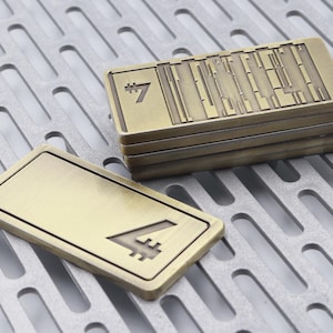 K&ouml;nnte beinhalten: Mehrere rechteckige Metallobjekte in Bronzefarbe mit strukturierter Oberfl&auml;che. Ein Objekt zeigt die Zahl "4". Die gestapelten Objekte haben die Zahl "7" und vertikale Linien. Die Objekte befinden sich auf einer perforierten Metalloberfl&auml;che.