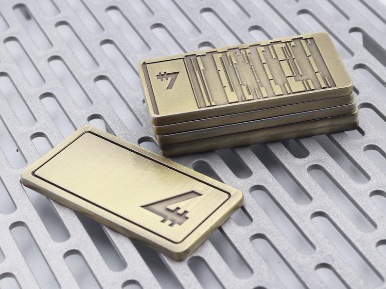 K&ouml;nnte beinhalten: Mehrere rechteckige Metallobjekte in Bronzefarbe mit strukturierter Oberfl&auml;che. Ein Objekt zeigt die Zahl "4". Die gestapelten Objekte haben die Zahl "7" und vertikale Linien. Die Objekte befinden sich auf einer perforierten Metalloberfl&auml;che.