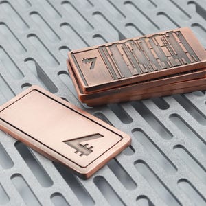 K&ouml;nnte beinhalten: Kupferfarbene rechteckige Metallobjekte mit erhabenem Design und der Zahl 7. Ein Objekt ist separat, w&auml;hrend andere gestapelt sind. Die Objekte haben eine strukturierte Oberfl&auml;che und werden auf einer grauen, gemusterten Oberfl&auml;che pr&auml;sentiert.