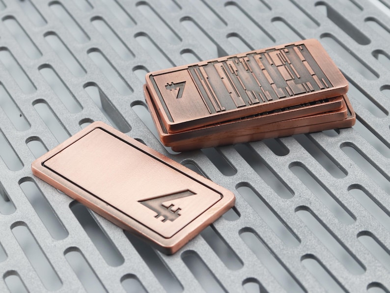 K&ouml;nnte beinhalten: Kupferfarbene rechteckige Metallobjekte mit erhabenem Design und der Zahl 7. Ein Objekt ist separat, w&auml;hrend andere gestapelt sind. Die Objekte haben eine strukturierte Oberfl&auml;che und werden auf einer grauen, gemusterten Oberfl&auml;che pr&auml;sentiert.