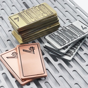 K&ouml;nnte beinhalten: Rechteckige Metallobjekte in Gold-, Silber- und Kupfert&ouml;nen, jeweils mit einer stilisierten "7" und geometrischen Designs. Die Objekte sind auf einer perforierten Metalloberfl&auml;che gestapelt, was auf ein Sammel- oder Dekorationsthema hindeutet.