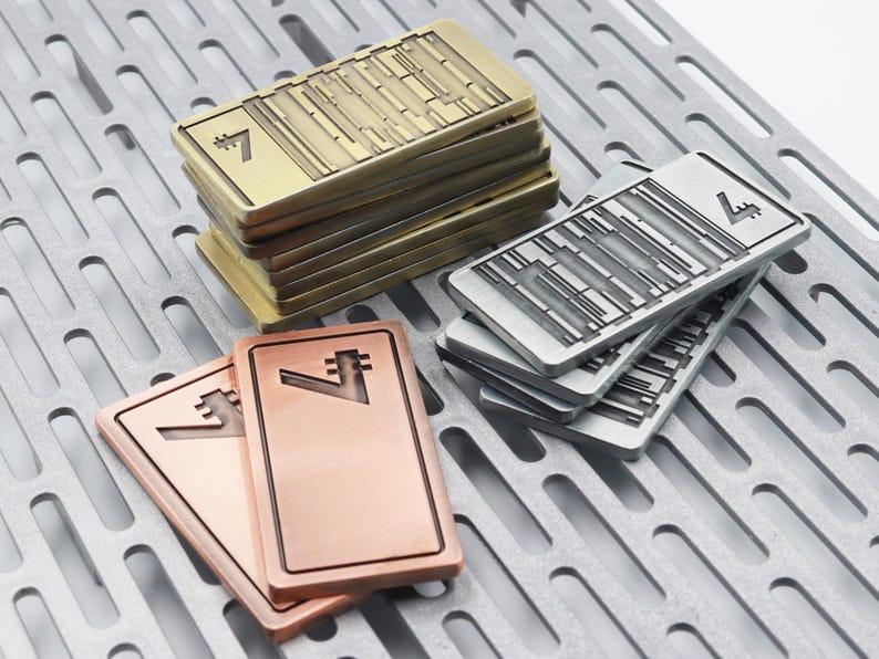 K&ouml;nnte beinhalten: Rechteckige Metallobjekte in Gold-, Silber- und Kupfert&ouml;nen, jeweils mit einer stilisierten "7" und geometrischen Designs. Die Objekte sind auf einer perforierten Metalloberfl&auml;che gestapelt, was auf ein Sammel- oder Dekorationsthema hindeutet.