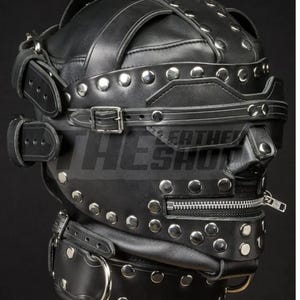 Puede incluir: Capucha de cuero negro con remaches plateados y cierre de cremallera en la boca. La capucha presenta múltiples correas, hebillas y un collarín con un gran anillo de metal. El texto "LEATHER SHOP" es visible.