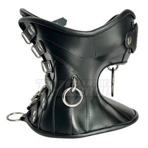 May include: Black leather neck collar with multiple silver-tone buckles and rings. The collar has a contoured design with a wide base and a narrower top, featuring a series of vertical seams for added detail. A metal ring is attached to the front.