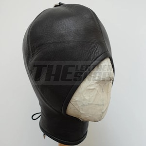 Puede incluir: Capucha de cuero negro con la parte superior redondeada y una correa para la barbilla. La capucha tiene aberturas para los ojos y la boca. El texto "THE LEATHER SHOP" es visible en el lateral de la capucha.