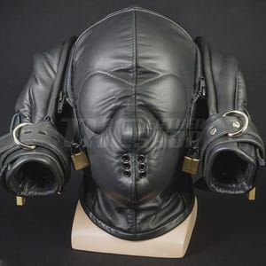 May include: Black leather hood with eye and nose cutouts, zippers, and attached wrist restraints. The hood features a series of small, decorative rivets and is secured with padlocks. The product is displayed on a mannequin head.