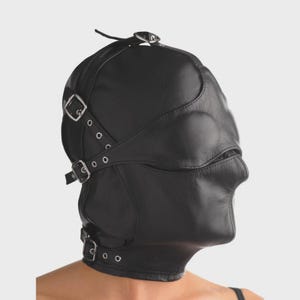 Puede incluir: Capucha de cuero negro con aberturas para ojos y boca. La capucha tiene correas ajustables con hebillas y remaches metálicos. El diseño cubre toda la cabeza y el cuello.