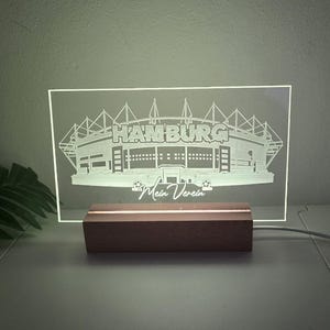 Könnte beinhalten: Ein beleuchtetes Schild mit dem Design des Hamburger Stadions, mit dem Wort "HAMBURG" und dem Satz "Mein Verein" in das klare Acryl geätzt. Das Schild ist auf einem Holzsockel montiert.