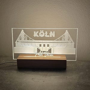 Könnte beinhalten: Ein beleuchtetes Schild mit der Umrisszeichnung eines Stadions und dem Wort "KÖLN" darüber. Das Schild steht auf einem Holzsockel mit den Worten "Mein Verein" darunter. Es leuchtet sanft weiß.