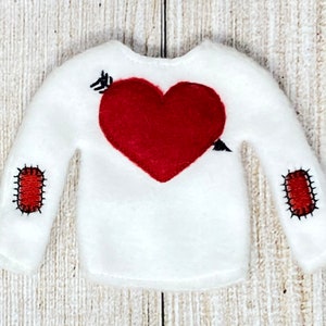 May include: White long-sleeved shirt with a red felt heart and arrow design on the front. The shirt has red felt patches on the elbows with black stitching.