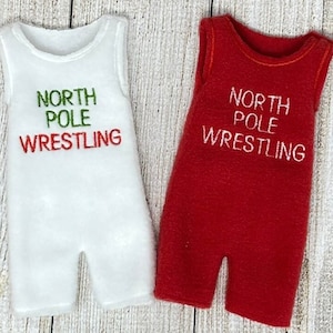 Puede incluir: Dos peleles sin mangas para un bebé o un niño pequeño. Uno es blanco con bordado verde y rojo que dice "North Pole Wrestling". El otro es rojo con bordado blanco que dice "North Pole Wrestling".