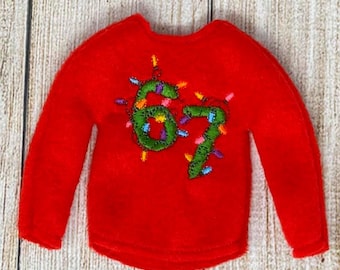 May include: A small, red sweater with long sleeves. The sweater features the embroidered number "67" in green, decorated with colorful Christmas lights. The garment is designed for a doll or small figure, perfect for holiday-themed displays.