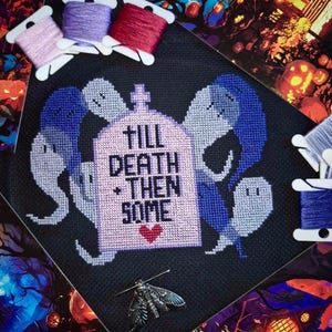 May include: Cross-stitch pattern featuring a tombstone with the text "Till Death + Then Some" and a heart. The tombstone is surrounded by four ghosts in shades of blue and purple. The pattern is framed and displayed on a black background.