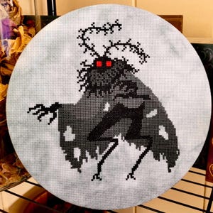 May include: A black and white cross-stitch embroidery of a moth with red eyes on a grey background.