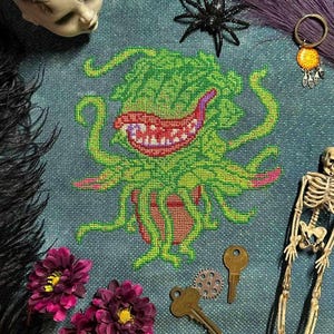 May include: A cross-stitch pattern of a green, toothy plant monster with pink tentacles, set against a blue background. The monster is in a red pot and has a toothy grin.